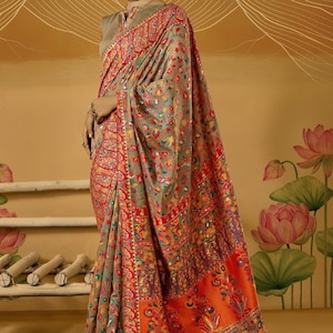 May include: A woman draped in a vibrant saree, showcasing detailed floral and paisley designs. The saree combines red, orange, and grey tones, with elaborate embroidery. The backdrop features a floral motif.