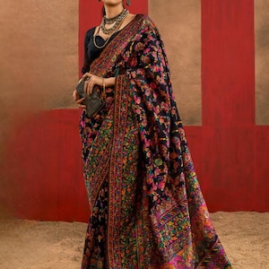 May include: A black sari with a vibrant floral pattern in shades of pink, green, orange, and yellow. The sari is draped over a person, showcasing the intricate embroidery and design. The background is a mix of red and tan.