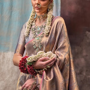 May include: A woman wearing a silver and gold saree with intricate embroidery. She is adorned with traditional Indian jewelry, including a nose ring, earrings, a necklace, and bangles. She is holding a garland of white flowers.