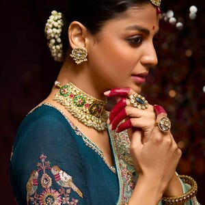 Op de afbeelding: Een vrouw draagt een tealblauwe blouse met ingewikkelde borduursels en een groen-gouden ketting. Ze draagt ook een zilveren armband en verschillende ringen aan haar vingers.