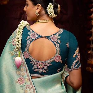 Op de afbeelding: Een tealblauwe blouse met een ronde uitsnijding in de rug en bloemenborduurwerk. De blouse wordt gedragen met een lichtblauwe sari met bloemenborduurwerk langs de zoom.