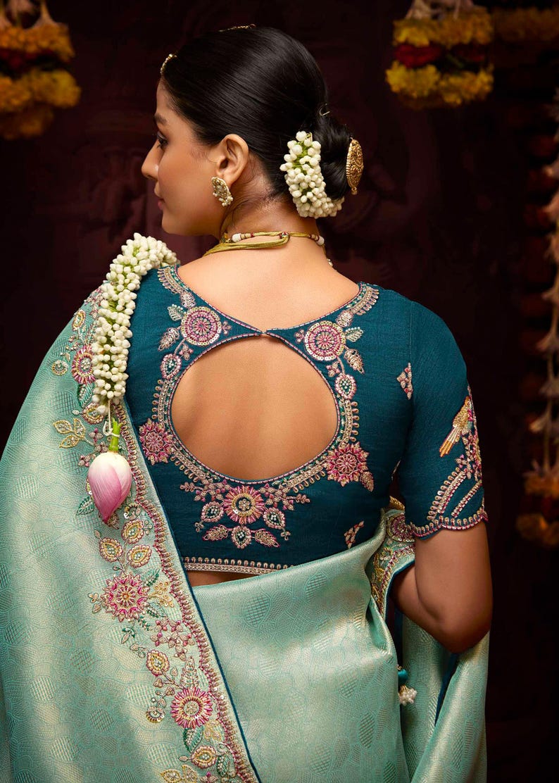 Op de afbeelding: Een tealblauwe blouse met een ronde uitsnijding in de rug en bloemenborduurwerk. De blouse wordt gedragen met een lichtblauwe sari met bloemenborduurwerk langs de zoom.