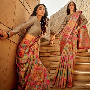 May include: A woman wearing a long-sleeved blouse and a colourful patterned saree. The saree has a floral and geometric design with shades of pink, blue, green, and gold. The woman is standing on a set of stairs.