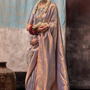 May include: A woman wearing a silver and gold saree with a pink border. She is wearing a floral garland and traditional jewelry.