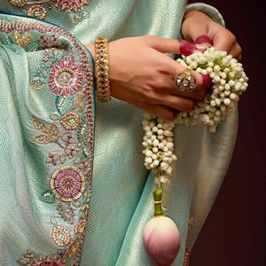 Op de afbeelding: Een lichtblauwe sari met ingewikkelde bloemenborduursels in roze en wit. De sari heeft een gouden rand met een textuurpatroon. De sari is over de arm van een vrouw gedrapeerd, en ze houdt een krans van witte bloemen vast.