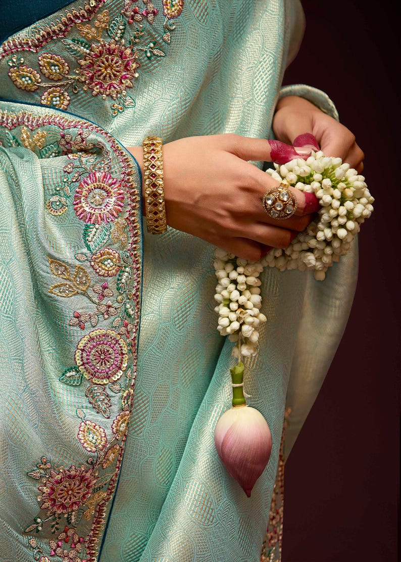 Op de afbeelding: Een lichtblauwe sari met ingewikkelde bloemenborduursels in roze en wit. De sari heeft een gouden rand met een textuurpatroon. De sari is over de arm van een vrouw gedrapeerd, en ze houdt een krans van witte bloemen vast.
