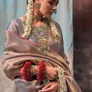 May include: A woman wearing a gold and purple silk saree with a pink border. She is adorned with traditional Indian jewelry, including a necklace, earrings, and a headpiece. She is also wearing a flower garland around her neck and wrist.
