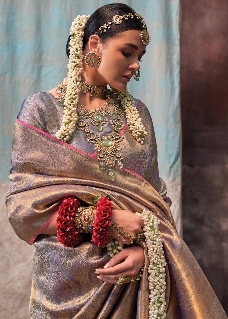 May include: A woman wearing a gold and purple silk saree with a pink border. She is adorned with traditional Indian jewelry, including a necklace, earrings, and a headpiece. She is also wearing a flower garland around her neck and wrist.