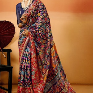 May include: A vibrant, patterned saree in shades of red, blue, and yellow. The fabric is adorned with intricate floral designs. The saree is worn with a navy blue blouse and layered pearl necklaces.