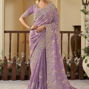 May include: A lavender silk saree with gold embroidery and sequins. The saree features a floral pattern and a border with intricate details.