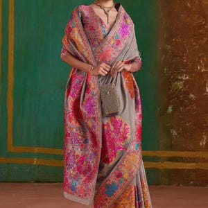 May include: A gray sari with intricate floral embroidery in pink, orange, and blue. The sari is draped over a person, with a matching shawl and a small, beaded purse. The background is a mix of green and brown.