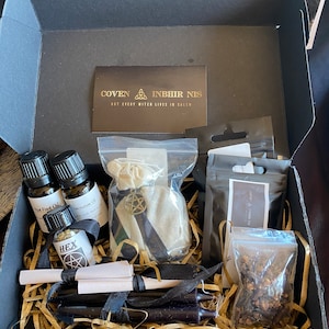 Könnte beinhalten: Eine offene schwarze Box gefüllt mit verschiedenen Gegenständen, darunter kleine Glasflaschen, schwarze Kerzen, ein kleiner Beutel mit einem Pentagramm-Anhänger und eine Karte mit dem Text "COVEN INBHIR NIS NOT EVERY WITCH LIVES IN SALEM". Die Box ist mit goldfarbenem Zierpapier ausgekleidet.