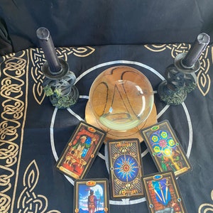 May include: A crystal ball on a wooden stand, with tarot cards and two candle holders with black candles. The cards are on a black cloth with gold Celtic knot designs and a white circle. The scene suggests divination.