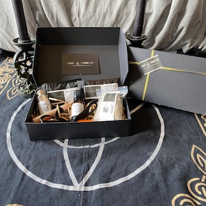 Könnte beinhalten: Eine schwarze Geschenkbox mit goldenen Akzenten, die verschiedene Artikel enthält. Im Inneren befinden sich kleine Flaschen, Päckchen und ein Fläschchen, alles auf Stroh gebettet. Die Box ist geöffnet und zeigt den Inhalt. Sie steht auf einem schwarzen Tuch mit weißem und goldenem keltischen Design. Zwei schwarze Kerzen flankieren die Box.
