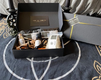 Peut inclure: Coffret cadeau noir avec bordure dorée, contenant divers articles. À l'intérieur, de petites bouteilles, des sachets et un flacon, le tout sur de la paille. Le coffret est ouvert, révélant le contenu, et repose sur un tissu noir avec un motif celtique blanc et or. Deux bougies noires encadrent le coffret.