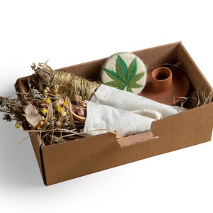 Peut inclure: Une boîte en carton remplie de produits de bain naturels. La boîte contient un bouquet d'herbes séchées, un tissu blanc, un savon en forme de feuille verte et un bougeoir en terre cuite.