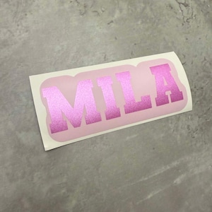 Könnte beinhalten: Rosaer Vinyl-Aufkleber mit dem Namen "MILA" in fetten, weißen Buchstaben.