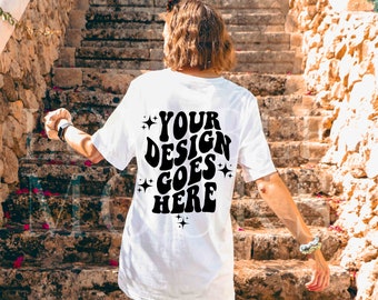 May include: A white short-sleeve t-shirt featuring black text on the back that reads "YOUR DESIGN GOES HERE" in a wavy, bold font, accented with small four-pointed star graphics. The shirt is worn with white sneakers and white socks.