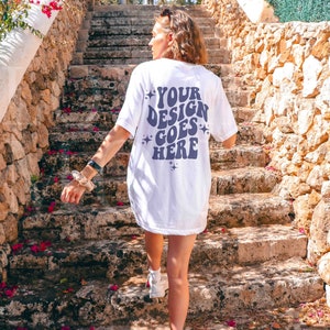 Op de afbeelding: Een wit T-shirt met een paars ontwerp op de achterkant met de tekst "YOUR DESIGN GOES HERE" met sterren rond de tekst. Het shirt wordt gedragen door iemand die een stenen trap oploopt.