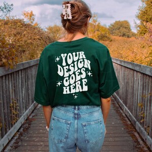Peut inclure: Une personne portant un t-shirt vert fonc&eacute; avec le texte "YOUR DESIGN GOES HERE" en blanc. Le t-shirt a une police de caract&egrave;res r&eacute;tro et des &eacute;toiles autour du texte. La personne porte un jean bleu et marche sur un pont en bois.