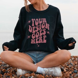 以下が含まれることがあります： 星が散りばめられた「YOUR DESIGN GOES HERE」というピンク色のデザインが施された黒いスウェットシャツ。砂浜の石の上に座っている人が着用しています。