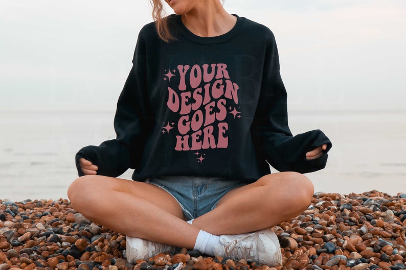 以下が含まれることがあります： 星が散りばめられた「YOUR DESIGN GOES HERE」というピンク色のデザインが施された黒いスウェットシャツ。砂浜の石の上に座っている人が着用しています。