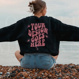 以下が含まれることがあります： キラキラのアクセントが付いたピンクの文字で「YOUR DESIGN GOES HERE」と書かれた黒いスウェットシャツ。ビーチに座っている人が着ています。