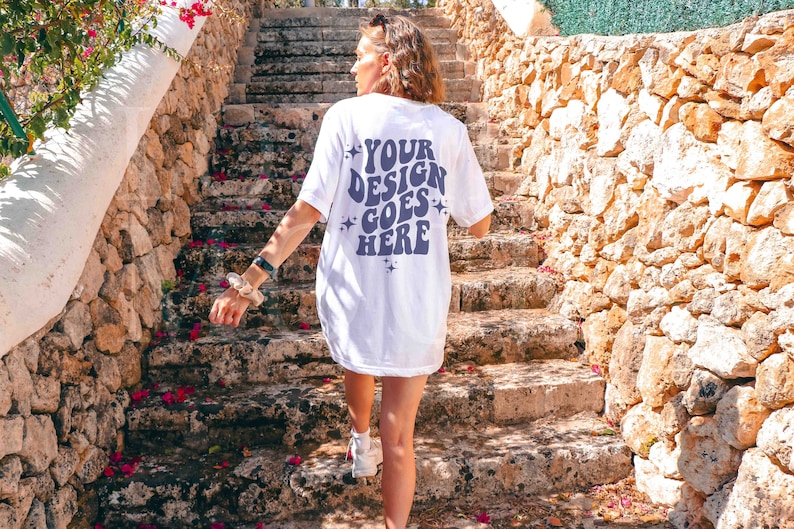 Op de afbeelding: Een wit T-shirt met een paars ontwerp op de achterkant met de tekst "YOUR DESIGN GOES HERE" met sterren rond de tekst. Het shirt wordt gedragen door iemand die een stenen trap oploopt.