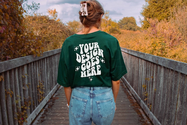 Peut inclure: Une personne portant un t-shirt vert fonc&eacute; avec le texte "YOUR DESIGN GOES HERE" en blanc. Le t-shirt a une police de caract&egrave;res r&eacute;tro et des &eacute;toiles autour du texte. La personne porte un jean bleu et marche sur un pont en bois.