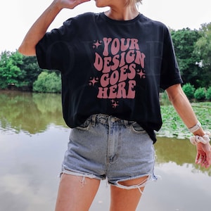 May include: A black t-shirt with the text "YOUR DESIGN GOES HERE" in pink lettering. The shirt is being worn by a person standing in front of a pond.