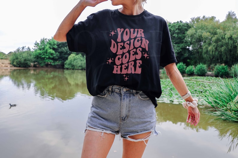 May include: A black t-shirt with the text "YOUR DESIGN GOES HERE" in pink lettering. The shirt is being worn by a person standing in front of a pond.