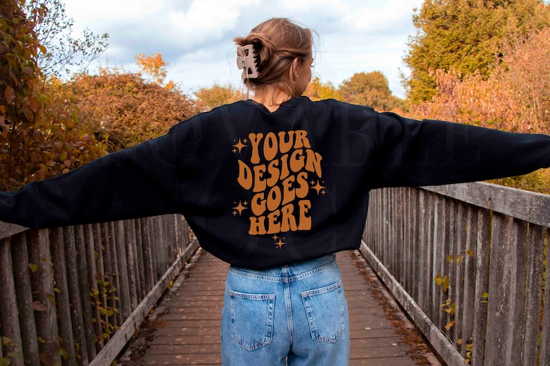 May include: A black sweatshirt with the text "YOUR DESIGN GOES HERE" in brown lettering on the back. The sweatshirt is being worn by a person standing on a wooden bridge.