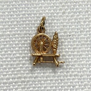 May include: A gold charm in the shape of a spinning wheel with a spindle. The charm is detailed and has a vintage look.