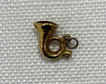 May include: A small, gold-colored French horn charm. The charm has a detailed design with a bell, tubing, and a loop for attaching to a necklace or bracelet. The charm is likely made of metal and has a polished finish.