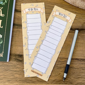 Puede incluir: Dos marcapáginas decorativos con fondo amarillo claro y estampado floral. Uno de los marcapáginas dice "TO BE READ" y el otro "WISH LIST". Un bolígrafo plateado con agarre negro descansa sobre una superficie de madera.