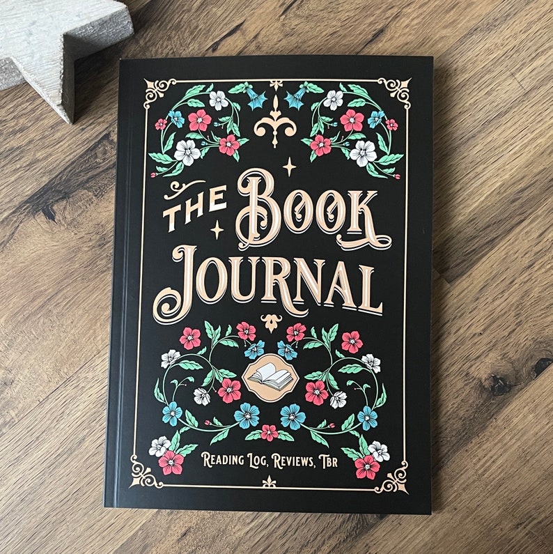 K&ouml;nnte beinhalten: Schwarzes Journal mit goldener Aufschrift "The Book Journal", umgeben von floralen Designs. Der Text "Reading Log, Reviews, TBR" befindet sich unten.