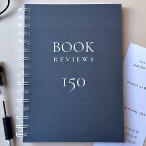 May include: A dark grey spiral-bound notebook with the words "BOOK REVIEWS" and the number "150" printed in white. A pen is visible on the left. A small piece of paper with text is on the right.
