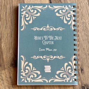Puede incluir: Un cuaderno encuadernado en espiral con una cubierta de color verde azulado con bordes decorativos de color crema y el texto "Here's To The Next Chapter" y "Love Mum xx". El dise&ntilde;o de la cubierta incluye una pila de libros y detalles ornamentales en las esquinas.