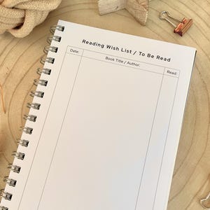 Puede incluir: Un cuaderno en espiral blanco con el texto "Reading Wish List / To Be Read". El cuaderno tiene secciones para la fecha, el t&iacute;tulo/autor del libro y una secci&oacute;n de lectura. El cuaderno est&aacute; abierto sobre una superficie de madera.