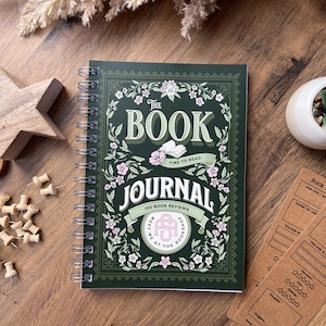 May include: A dark green spiral-bound book journal with the title "The Book Journal" and floral decorations. The image also shows a wooden star, push pins, a small succulent in a white pot, and two brown bookmarks with space for notes.