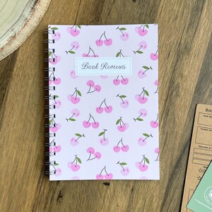 May include: A spiral-bound notebook with a light pink cover featuring a repeating pattern of pink cherries with green leaves. A white rectangular label in the center contains the black text Book Reviews in a cursive font style.