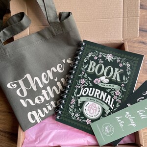 May include: An olive green tote bag with the text "There's nothing quit" next to a spiral-bound journal. The journal has a floral design and the words "The Book Journal" on the cover. Two bookmarks are also visible.