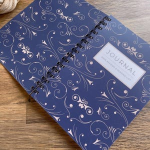 May include: An open spiral-bound journal with a navy blue cover and a cream floral pattern. The journal displays the text "JOURNAL" and "BOOK REVIEWS & READING LIST" within a rectangular frame. It rests on a wooden surface.