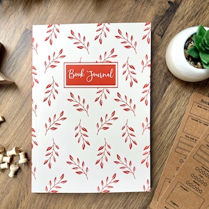 May include: A white book journal with a red floral pattern and the words "Book Journal" printed on the front cover.