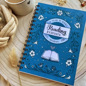 May include: A blue reading journal with a spiral binding, decorated with floral and leaf patterns. The cover displays the text "A Year in Books Reading Journal Log Reviews TBR" and an open book illustration. A cup of coffee is visible in the background.