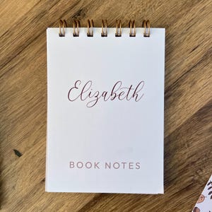 May include: A white notepad with a gold spiral binding, featuring the name "Elizabeth" in cursive and the text "BOOK NOTES". The notepad rests on a wooden surface, offering a simple, elegant design.