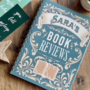 May include: A teal spiral-bound notebook with the words "Sara's Book Reviews" in a decorative font. The cover features an illustration of an open book and scrollwork. Additional cards and a wooden star are visible.