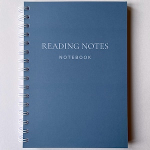 May include: A blue spiral-bound notebook with the text "READING NOTES NOTEBOOK" in white. The notebook has a minimalist design, ideal for journaling or note-taking. The spiral binding is white.