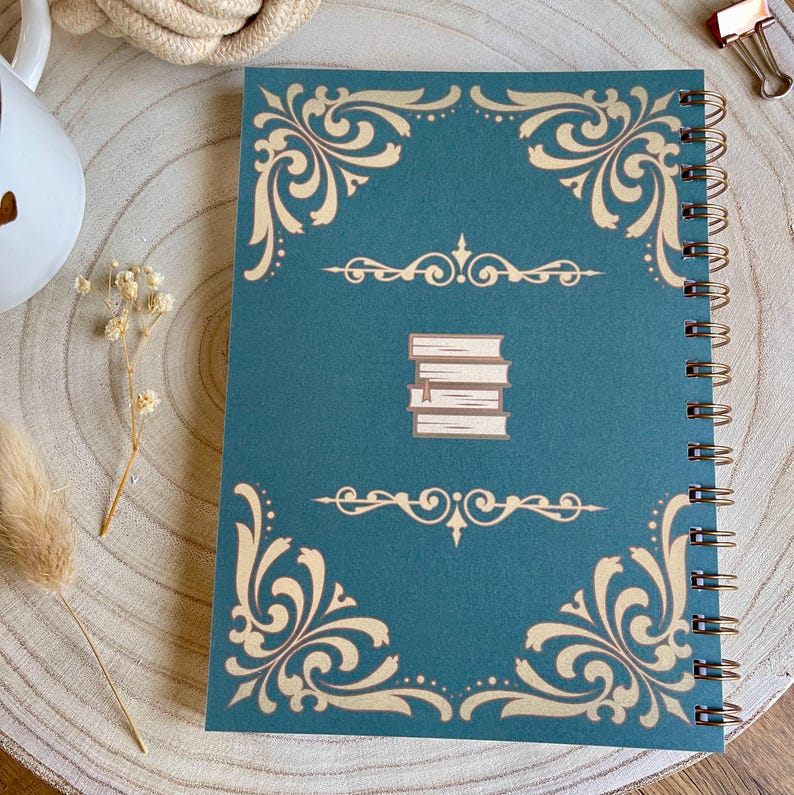 Puede incluir: Un cuaderno en espiral de color turquesa con dise&ntilde;os dorados de filigrana en las esquinas y bordes. La portada presenta una ilustraci&oacute;n de una pila de libros. El cuaderno est&aacute; abierto, revelando la encuadernaci&oacute;n en espiral. Sobre una superficie de madera.