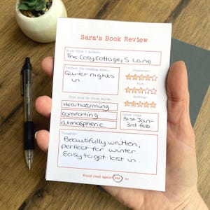 Könnte beinhalten: Eine Buchrezensionskarte mit dem Titel "Sara's Book Review" mit handschriftlichen Notizen. Die Karte enthält den Buchtitel "The Cosy Cottage, S. Lane" und die Wörter "Heartwarming, comforting, atmospheric". Die Karte hat auch einen Bewertungsbereich und die Lesedaten.